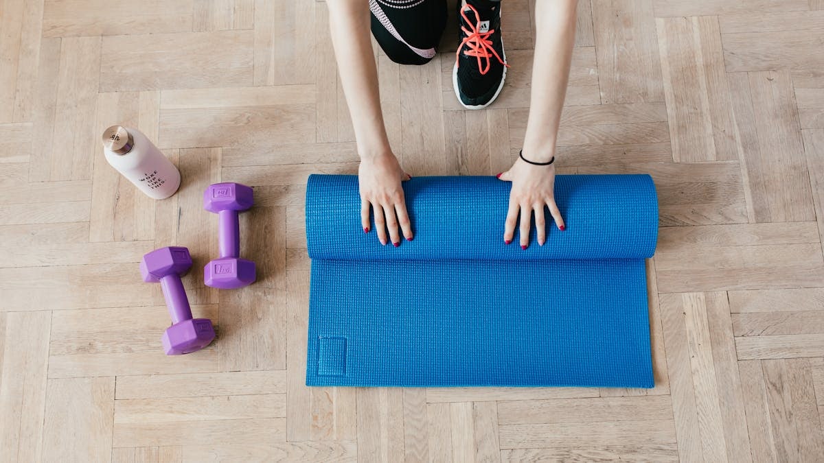 Top view of anonymous female athlete in black leggings and sneakers unfolding blue mat for exercising on floor near dumbbells and water bottle in modern fitness center